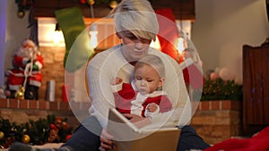 Positive young woman reading book for curios baby girl sitting in living room at fireplace. Front view portrait of happy