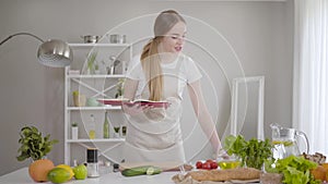 Positive young woman reading book of recipes and checking ingredients for dishes. Smiling Caucasian girl cooking dinner