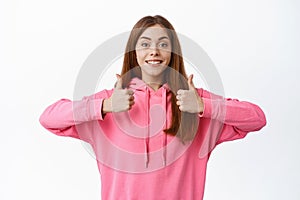 Positive young woman shows thumbs up, approve and like good product, agree or paise choice, stands against white