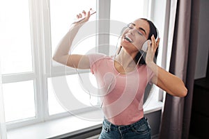 Positive young woman stand and listen to music through headphones in room. She dance and enjoy. Model smile. She hold