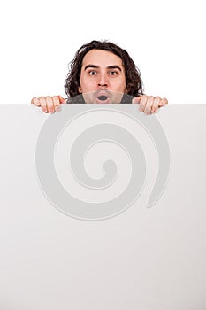Positively surprised young man, long curly hair style, staring with big eyes and mouth open, behind a big, blank announcement