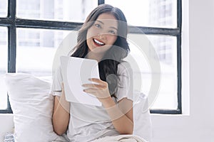 Positivity asian woman holding digital tablet sitting with pillow white bed beside window at home. Happy young female sitting