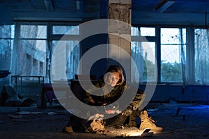 Post-apocalypse. a young woman sits in an abandoned building on the floor, takes out a gas burner for warming. concept