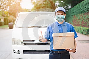 Postal delivery courier man thumbs up wearing protective face mask in front of cargo van delivering package holding box due to