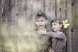 Postcard, stylized as vintage for the Victory Day. A boy in a military uniform and a girl in an old dress are standing near an old