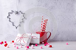 Postcard with  wrapped presents, heart, paper straws in red pitcher, pink and red pom poms against  grey  textured  wall.