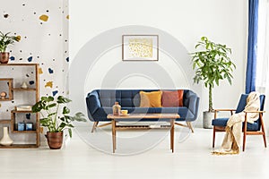 Poster above blue settee in white apartment interior with armchair, wooden table and plants. Real photo