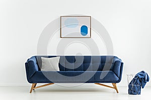 Poster above blue sofa with cushion in white living room interior with blanket in basket. Real photo