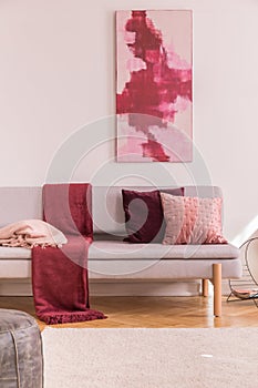Poster above grey settee with red blanket and cushions in bright living room interior. Real photo