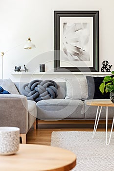 Poster above grey sofa with cushions in white living room interior with lamp and table. Real photo