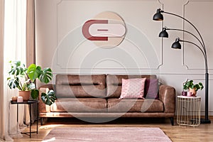 Poster above leather couch with pillows in white flat interior with plants and lamp