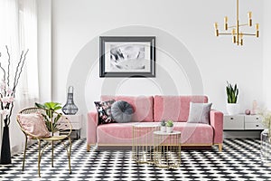 Poster above pink sofa in living room interior with gold armchair on checkered floor. Real photo