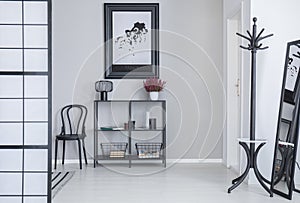 Poster above shelves with flowers and lamp in white simple hall interior with rack and black chair