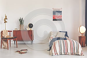 Poster above striped bed in white vintage bedroom interior with chair next to wooden cabinet. Real photo