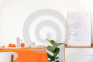 Poster on cabinet next to plant and table with orange cloth in white dining room interior
