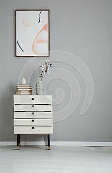 Poster on grey wall above white cabinet with books and flowers in simple flat interior. Real photo