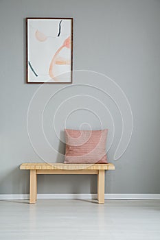 Poster on grey wall above wooden bench with pink pillow in simple apartment interior. Real photo