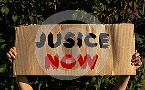 A poster with the words JUSICE NOW in the hands of a young girl on a background of green foliage