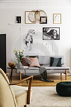 Posters above grey sofa with pillows in living room interior with flowers and armchair. Real photo