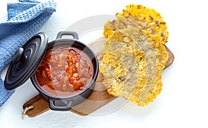 Pot with fried tomato in a black bowl accompanied by fried banana on a white background. Colombian cuisine concept. PatacÃ³n con