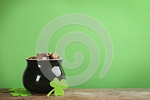 Pot of gold coins and clover leaves on wooden table against green background, space for text. St. Patrick`s Day celebration