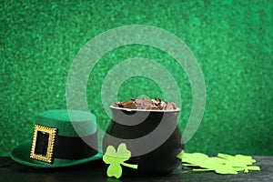 Pot of gold coins, hat and clover leaves on black stone table against green background, space for text. St. Patrick`s Day