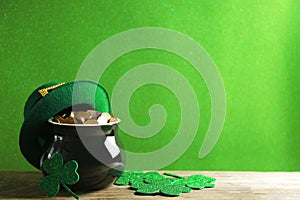 Pot of gold coins, hat and clover leaves on wooden table against green background, space for text. St. Patrick`s Day celebration