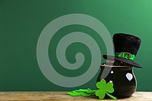 Pot of gold coins, hat and clover leaves on wooden table against green background, space for text. St. Patrick`s Day celebration