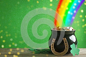 Pot with gold coins, horseshoe and clover leaves on wooden table against green background, space for text. St. Patrick`s Day