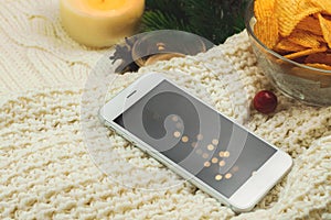 Potato chips in bowl and smartphone, cozy winter composition with garland bokeh, woolen sweater