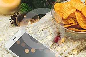 Potato chips in bowl and smartphone, cozy winter composition with garland bokeh, woolen sweater