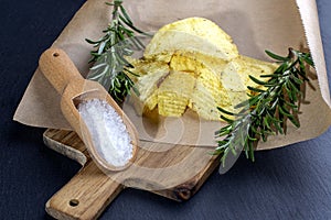 Potato chips with wooden spoon salt and rosematy on a wooden board. Fast food. Dark background