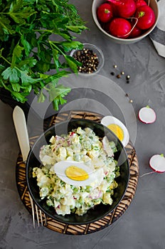 Potato salad with radish, eggs, green peas, sour cream and mayonnaise in a bowl on a board on a dark concrete background. Vertical