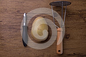 A potatoe with a tool to crushed potatoe and a peeler in the center of a wooden table