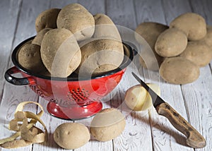 Potatoes in a drainer on a white wooden board on which there are other potatoes with one peeled potato and a knife