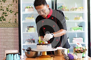 Potrait of asian male pastry chef in a black uniform decorating round vanila cake with melted chocolate and red cherry