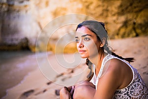 Potrait latin girl sitting on golden sand at beach near rocks and looking at ocean. Summer vocation