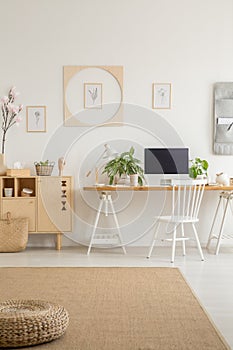 Pouf on carpet in white workspace interior with posters above desk with desktop computer. Real photo