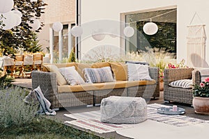 Pouf next to rattan couch and armchair on wooden terrace with flowers and lamps