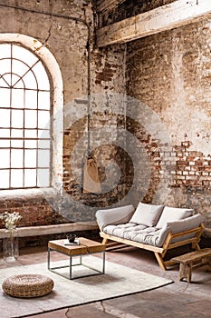 Pouf and table on rug in front of wooden sofa in spacious loft interior with window photo