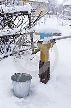 Pour clean drinking water into a bucket using a water pump, on a frosty winter evening, against a blurred background of the garden