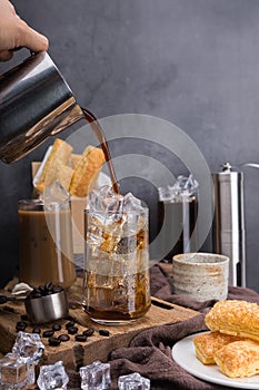 Pour coffee of americano coffee into a transparent glass with ice. and roasted coffee beans, sugar crispy pie, and hot tea on a bl