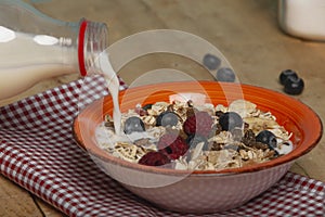 Pour the milk on the plate with oat flakes, dried fruits with blueberries and raspberries on wooden background. Healthy breakfast