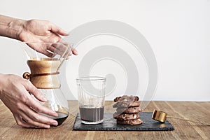Pour over coffee brewing method Chemex, woman hands hold a glass bowl, still life with brownie cookies on wooden table.