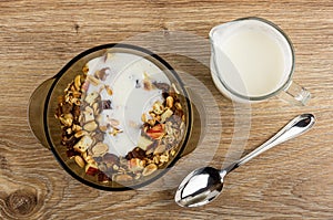 Poured yogurt granola in brown bowl, pitcher with yogurt, spoon on wooden table. Top view