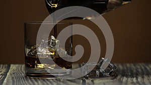 Pouring alcohol drink whiskey, cognac into glass with ice. Dark background.