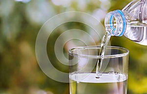 Pouring of clear drink water from bottle into the glass on blurred green nature background with copy space for text