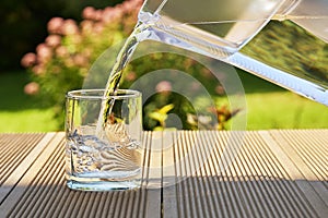 Pouring clear filtered water from a water filtration jug into a glass in green summer garden in a sunny summer day