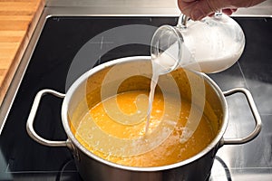 Pouring coconut milk or cream in a vegetable soup from red kuri squash or Hokkaido pumpkin in a steel pot on a black stove,