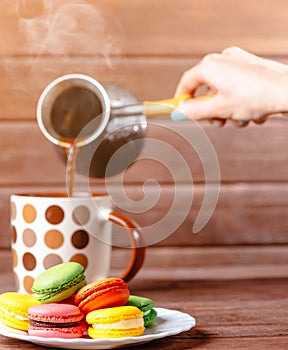 Pouring coffee from cezve to cup near macarons dessert.